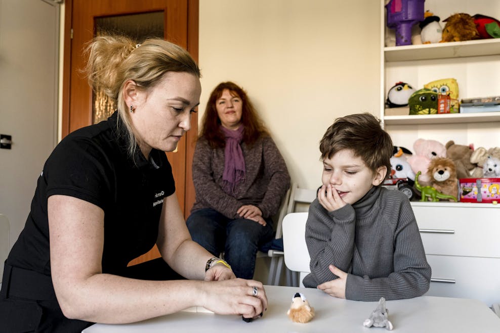 Een blonde vrouw zit aan tafel met een jongetje met donker haar en een grijze trui. Achter hen zit nog een vrouw, de moeder van het jongetje.