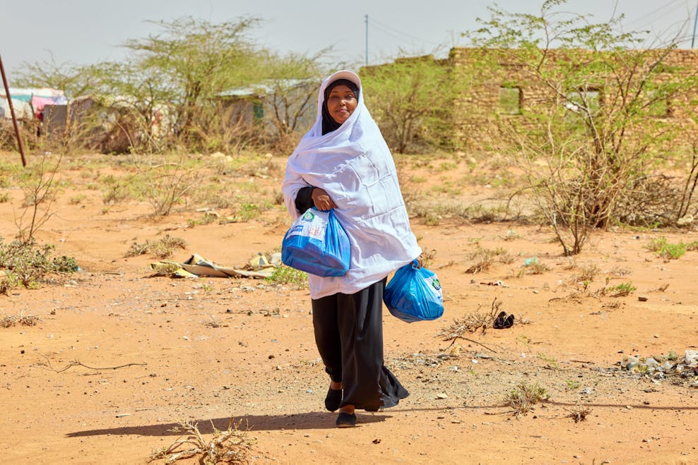 Menstruatie stopt niet voor noodsituaties Een zwarte vrouw met een witte jas die ook haar hoofd bedekt. In haar handen heeft ze twee blauwe tasjes met menstruatiekits.