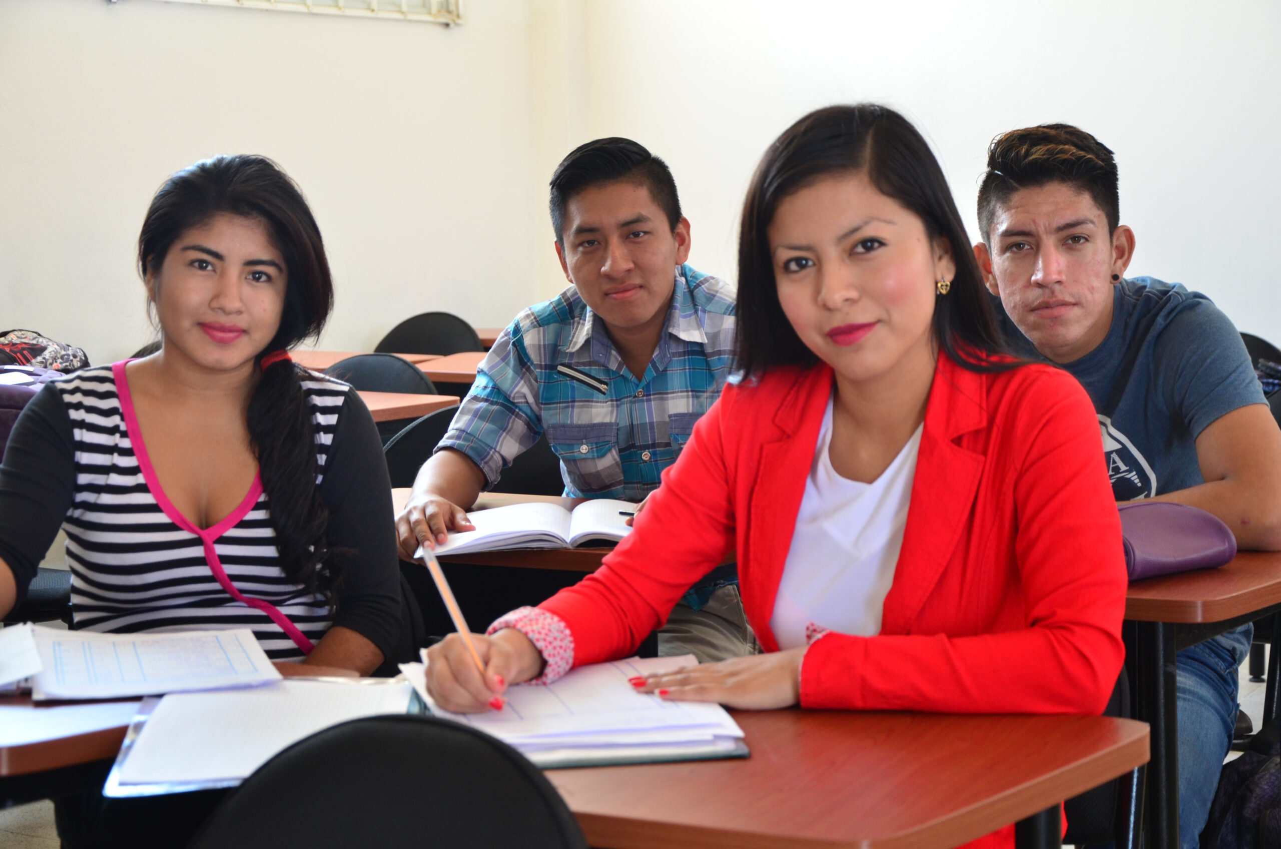 Vier jongeren zitten in een klaslokaal. Op de voorgrond zit Estefania in een rode blazer en een wit t-shirt. Ze heeft rode lippenstift op. Naast haar zit een ander meisje en achter haar twee jongens.