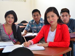 Vier jongeren zitten in een klaslokaal. Op de voorgrond zit Estefania in een rode blazer en een wit t-shirt. Ze heeft rode lippenstift op. Naast haar zit een ander meisje en achter haar twee jongens.