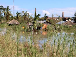 Een veld in Mozambique is overstroomd als gevolg van orkaan Freddy. Op de voorgrond zie je gras, dan water en op de achtergrond huizen.