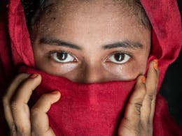 Een meisje kijkt recht in de camera. Ze draagt een rode hoofddoek en verbergt hiermee ook haar neus en mond. Ze houdt haar handen tegen haar gezicht.