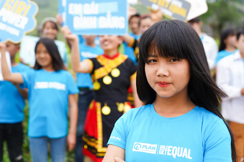 Een Vietnamees meisje in een blauw t-shirt kijkt recht in de camera. Achter haar staan andere meisjes die protesteren voor meisjesrechten.