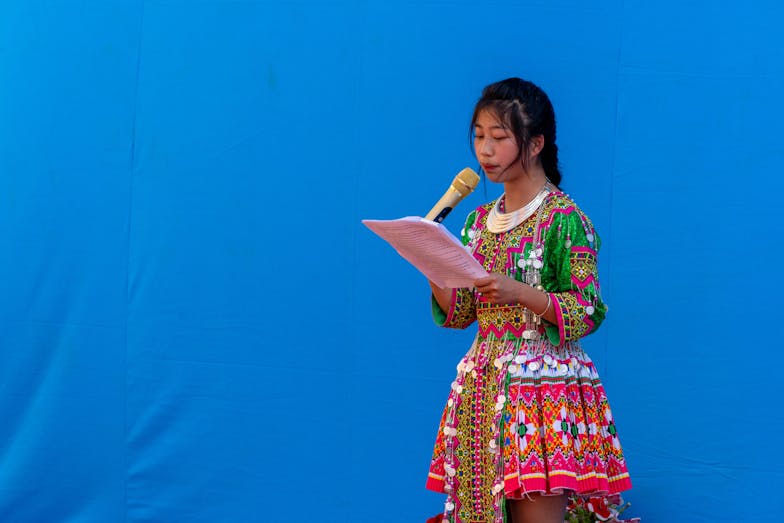 Een meisje in een kleurrijke jurk staat voor een blauwe achtergrond. Ze houdt een gouden microfoon vast en leest voor.