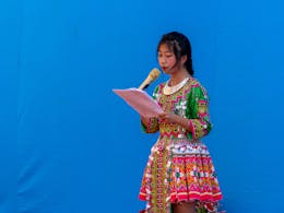 Een meisje in een kleurrijke jurk staat voor een blauwe achtergrond. Ze houdt een gouden microfoon vast en leest voor.