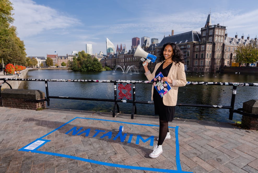Natanim (20) overhandigt Wereldmeisjesdagrapport aan ministerie en Kamerleden Een meisje staat voor de hofvijver in Den Haag. Ze staat in een vlak dat is gemarkeerd met blauwe tape, in het vak staat haar naam: Natanim. In haar handen houdt ze een microfoon en een rapport.