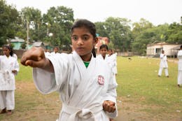 Masuma (12) uit Bangladesh staat in een karatepose met haar hand naar voren en haar vuist gebald. Ze draagt een wit karatepak.