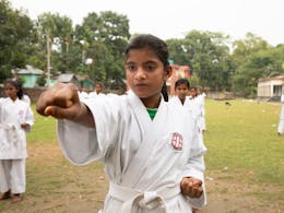 Masuma (12) uit Bangladesh staat in een karatepose met haar hand naar voren en haar vuist gebald. Ze draagt een wit karatepak.