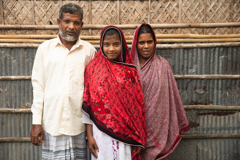 Een gezin van drie kijkt in de camera. Links staat de vader van Masuma, in het midden Masuma met een rode hoofddoek op, en rechts haar moeder met een paarse hoofddoek op.