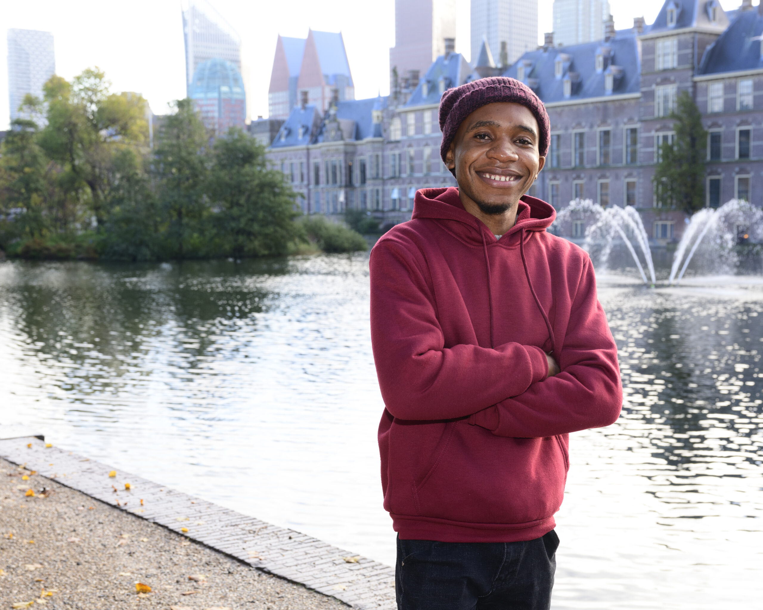 Een jongen in een rode hoodie en met een rode muts op staat voor de Hofvijver in Den Haag. Hij lacht naar de camera.