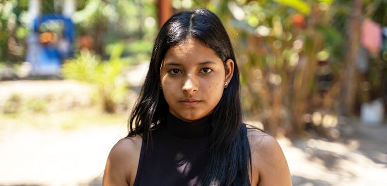 Carmen (16) kijkt recht in de camera. Ze heeft lang zwart haar en draagt een zwarte top.