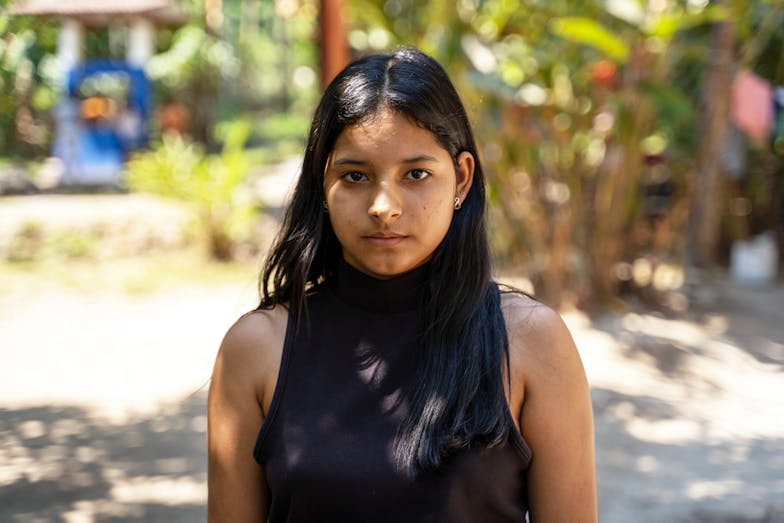 Carmen moest alles achterlaten: “Voor meisjes is vluchten extra gevaarlijk” Carmen (16) kijkt recht in de camera. Ze heeft lang zwart haar en draagt een zwarte top.