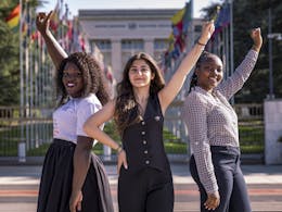 Drie meisjes staan voor het gebouw van de Verenigde Naties in Genève. Ze steken alledrie een hand in de lucht.