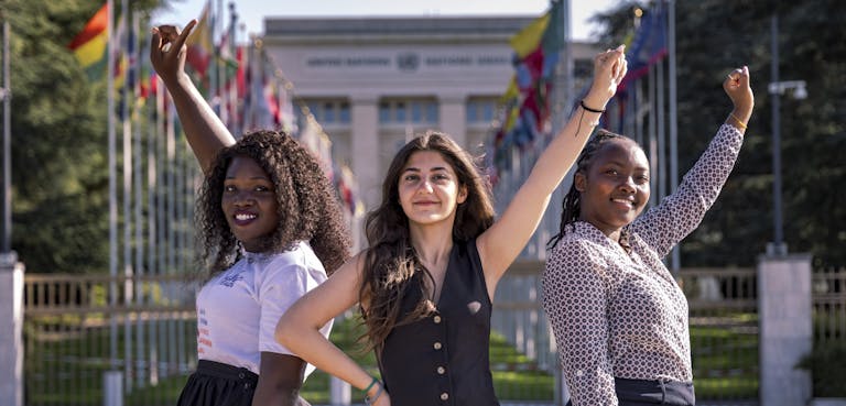 Drie meisjes staan voor het gebouw van de Verenigde Naties in Genève. Ze steken alledrie een hand in de lucht.