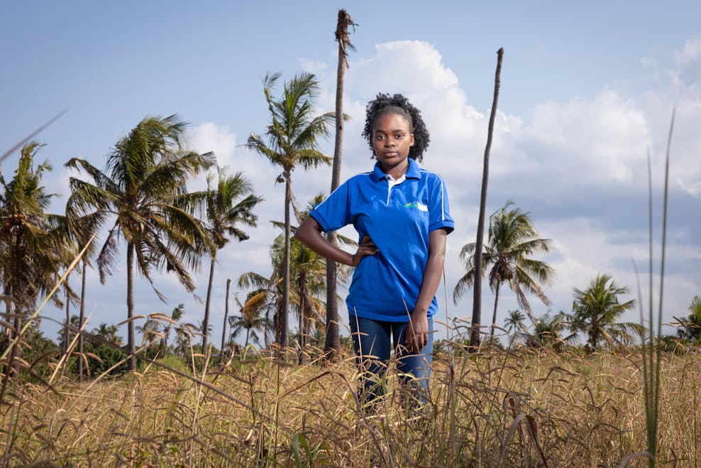 Een meisje in een blauw shirt staat in een veld met palmbomen op de achtrgrond. Ze houdt een hand in haar zij en kijkt stoer in de camera.