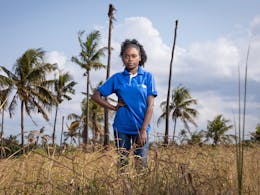 Een meisje in een blauw shirt staat in een veld met palmbomen op de achtrgrond. Ze houdt een hand in haar zij en kijkt stoer in de camera.