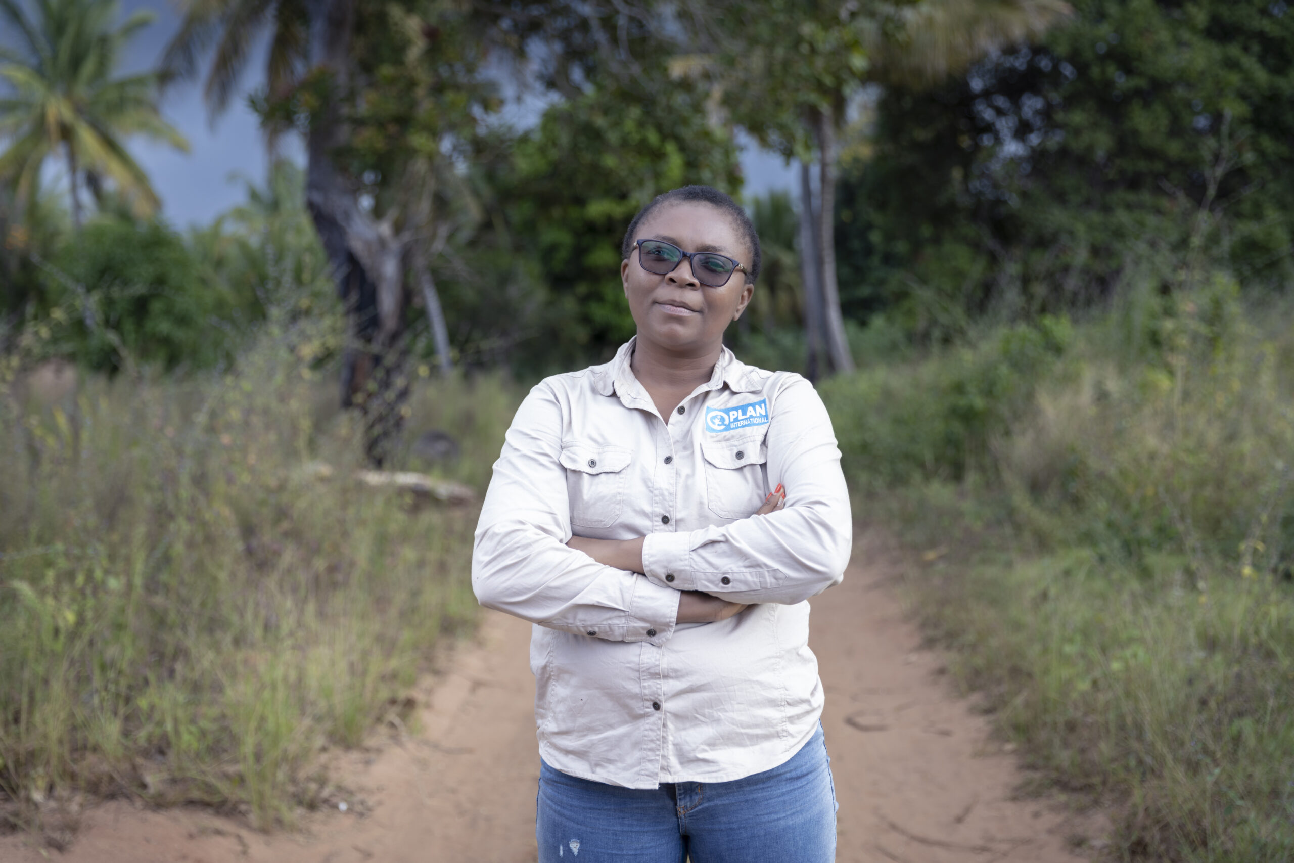 Een zwarte vrouw kijkt zelfverzekerd in de camera. Ze draagt een zonnebril en een beige shirt met een blauw Plan International-logo.