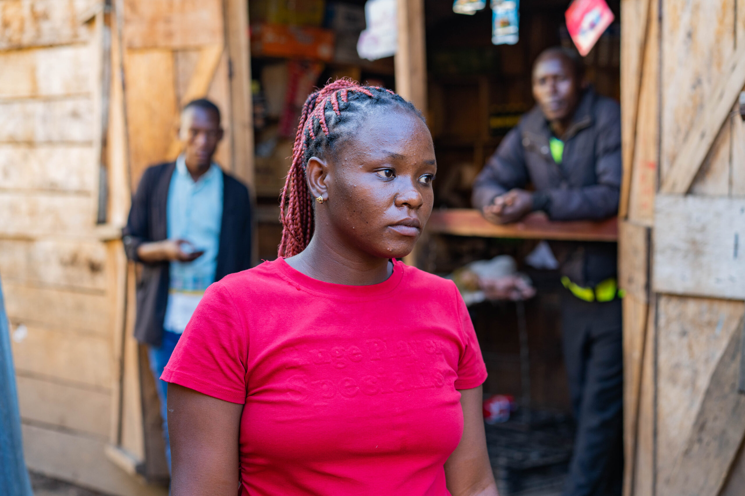 Een meisje in een rood t-shirt loopt door een straat. Achter haar staan twee mannen die niet scherp in beeld zijn. Ze kijken haar na.