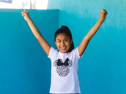 Een Peruaans meisje kijkt lachend in de camera terwijl ze haar handen in de lucht houdt. Ze draagt een wit t-shirt met Minnie Mouse erop.