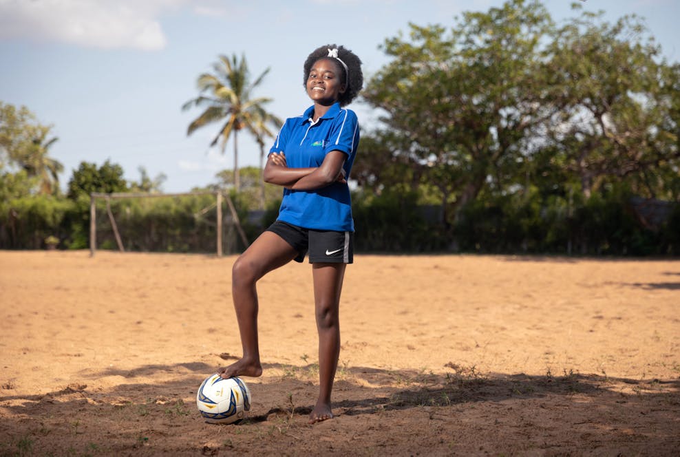 Kindhuwelijken voorkomen is een teamsport Een meisje uit Mozambique staat op een zandvlakte. Ze draagt een blauw t-shirt, onder haar voet houdt ze een voetbal.