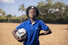 Een meisje uit Mozambique kijkt stoer in de camera. Ze draagt een blauw t-shirt en heeft een voetbal onder haar arm.