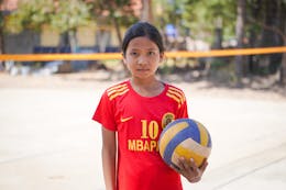 Een meisje met zwart haar en een rood voetbalshirt kijkt recht in de camera. In haar hand houdt ze een volleybal.