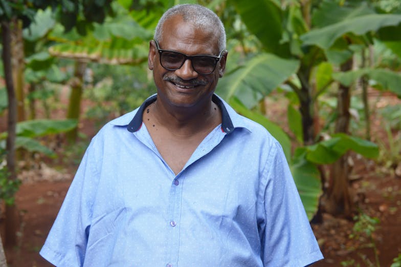 Een oudere man kijkt lachend in de camera. Hij draagt een donkere zonnebril en een blauw overhemd met korte mouwen.