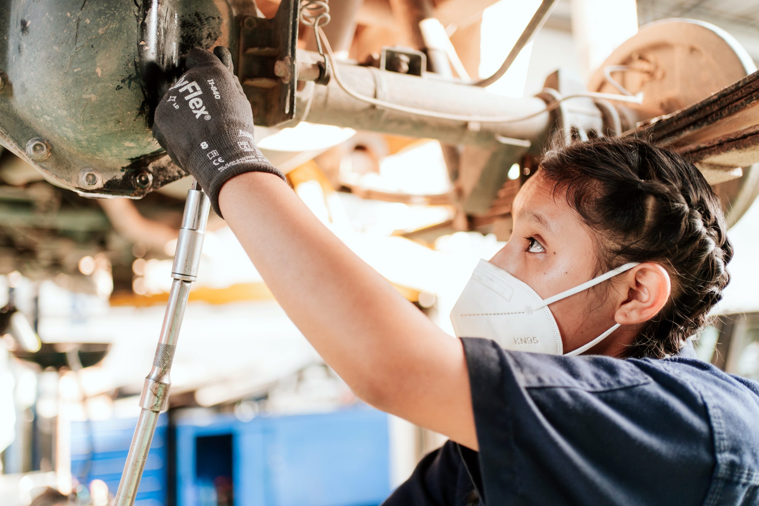 Een meisje staat met een mondkapje op onder het onderstel van een auto die ze repareert.