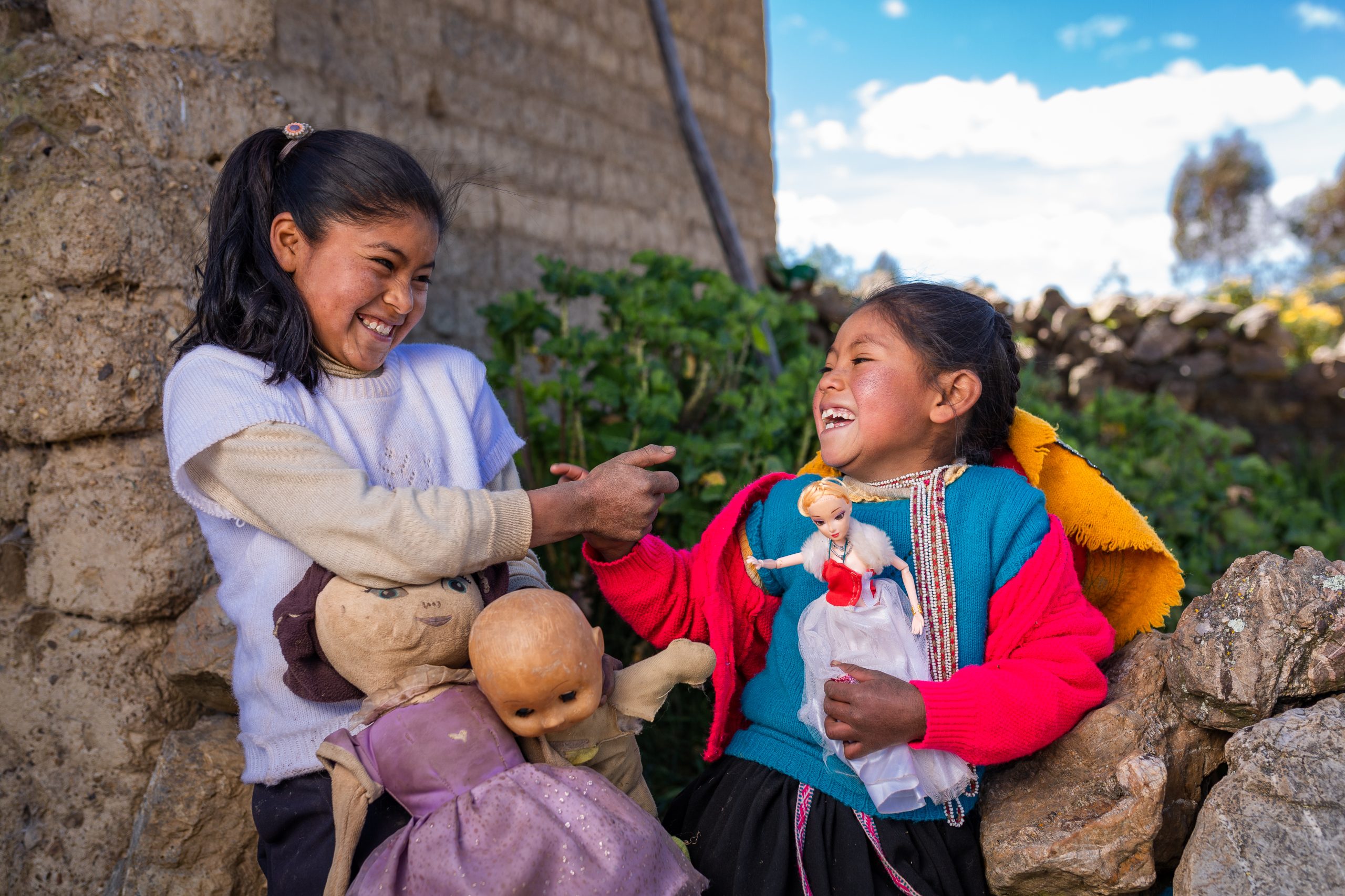 Twee lachende kinderen uit Cusco wijzen naar elkaar. In hun hand houden ze poppen om mee te spelen.