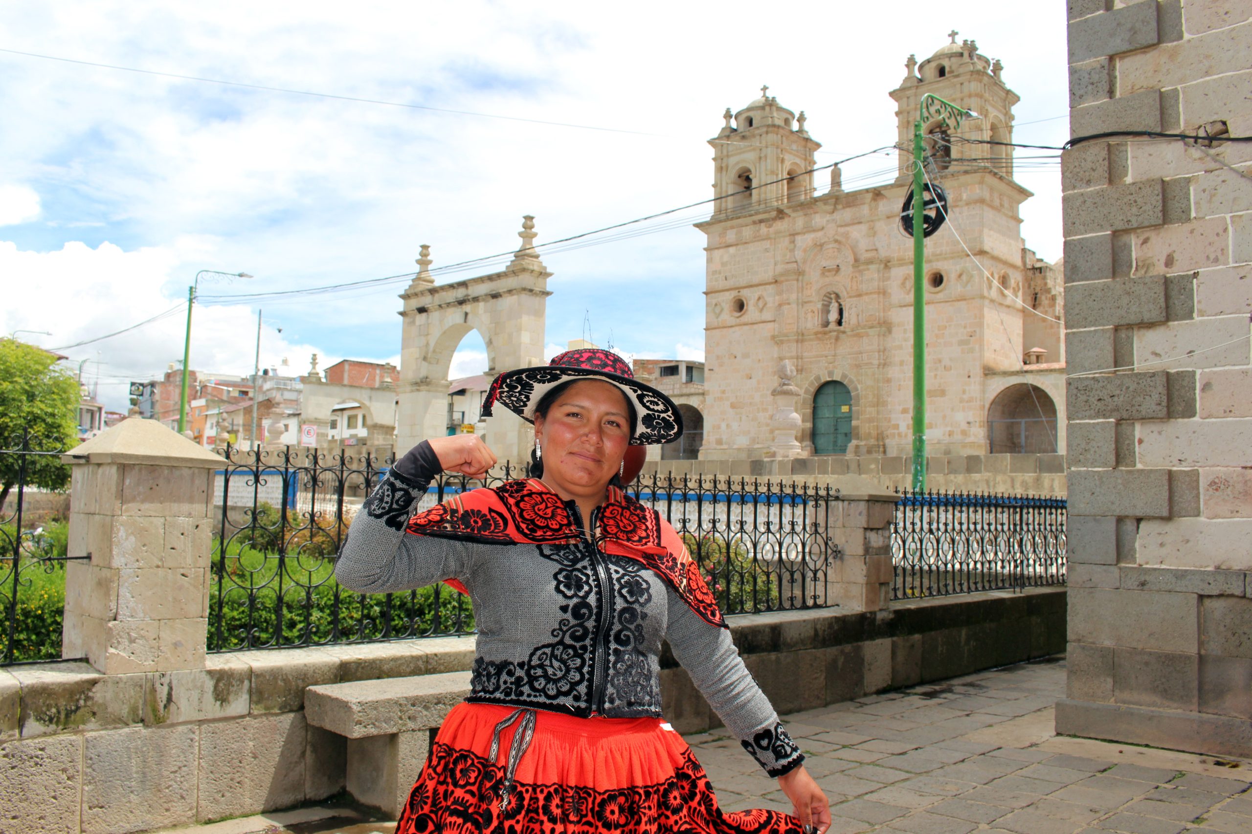 Een vrouw in traditionele kleding staat voor witte, stenen gebouwen. Haar arm houdt ze gebogen in de lucht, om haar spierballen te laten zien.