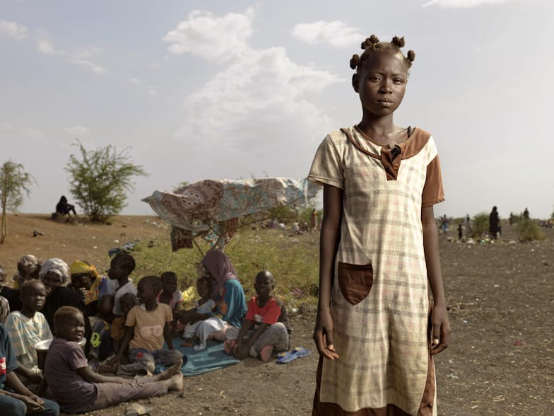 Jacklyn (12) uit Sudan kijkt boos de camera in. Ze draagt een geblokte jurk. Achter haar zit een groep mensen op de grond.